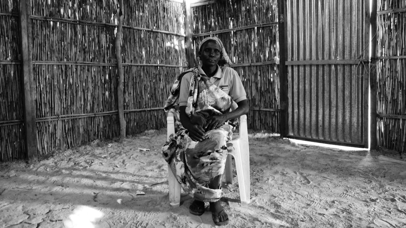 A person sits on a plastic chair inside a hut made of vertical wooden sticks and a corrugated metal door, holding a small child wrapped in a patterned cloth